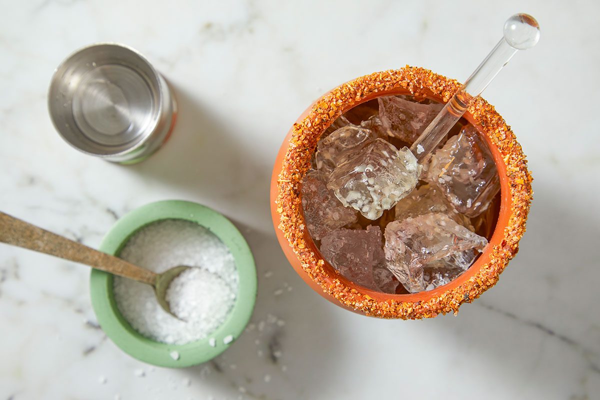 Ice cubes are added to a Tajín-rimmed clay glass alongside salt and bar tools to begin building the cocktail.