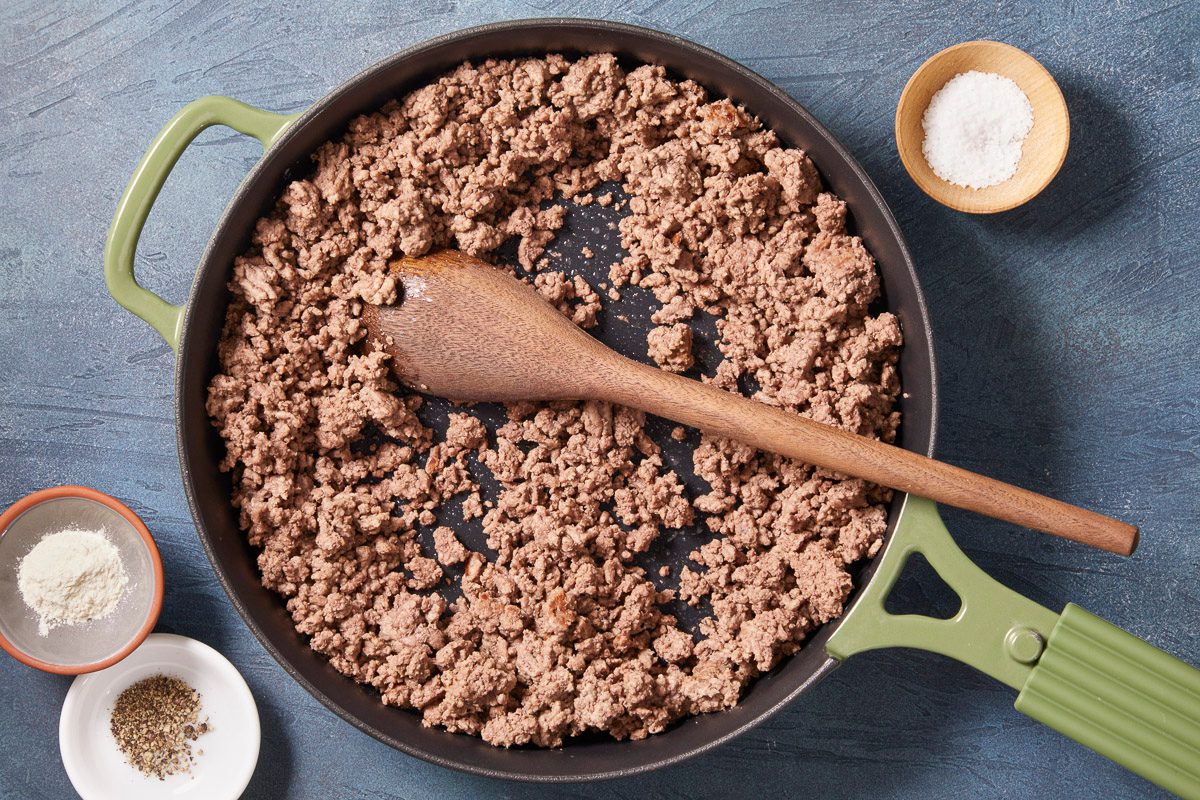 A horizontal overhead image shows browned ground beef cooking in a skillet with a wooden spoon and small bowls of seasoning nearby.