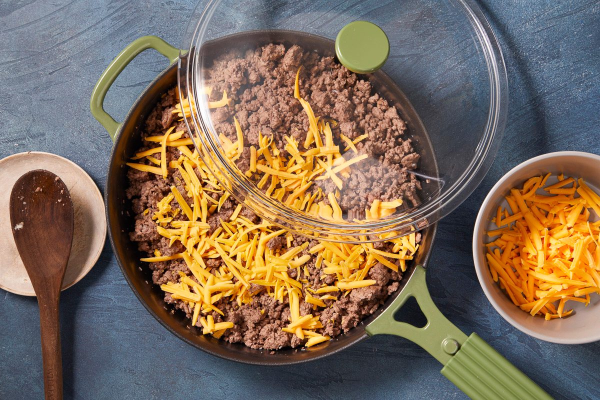 A horizontal overhead image shows shredded cheddar cheese being added to cooked ground beef in a skillet with a lid partially covering the pan.