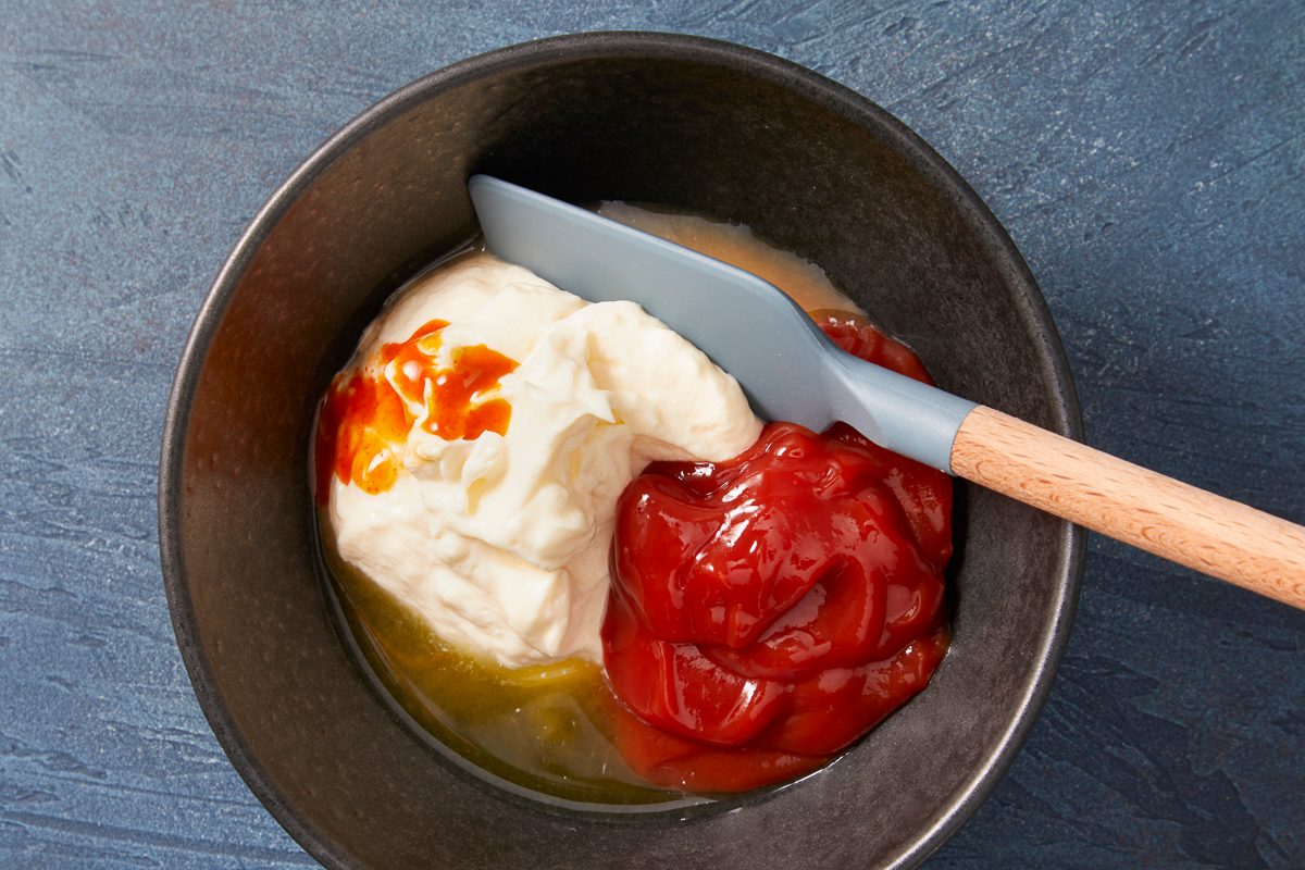 A horizontal overhead image shows a bowl containing mayonnaise, ketchup, and hot sauce being mixed together into a creamy sauce.