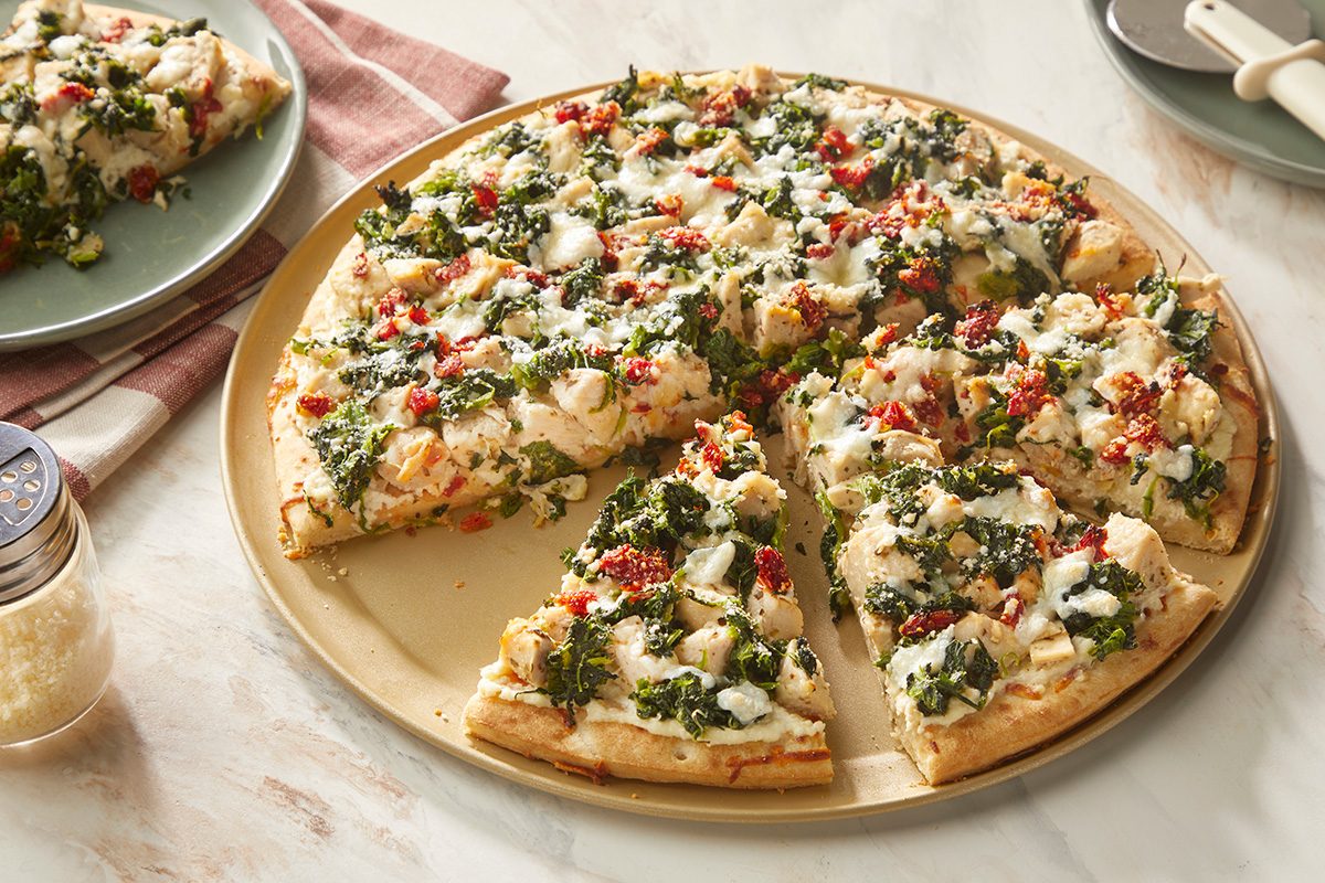 A cooked pizza topped with spinach, chunks of chicken, diced tomatoes, and melted cheese sits on a round baking tray. One slice has been removed and placed on a plate nearby.