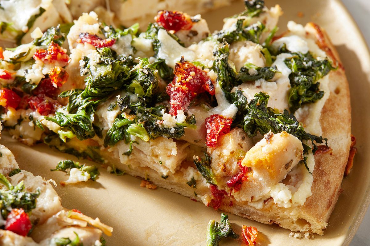 A close-up of a pizza slice topped with pieces of chicken, chopped spinach, sun-dried tomatoes, and melted cheese on a beige plate.