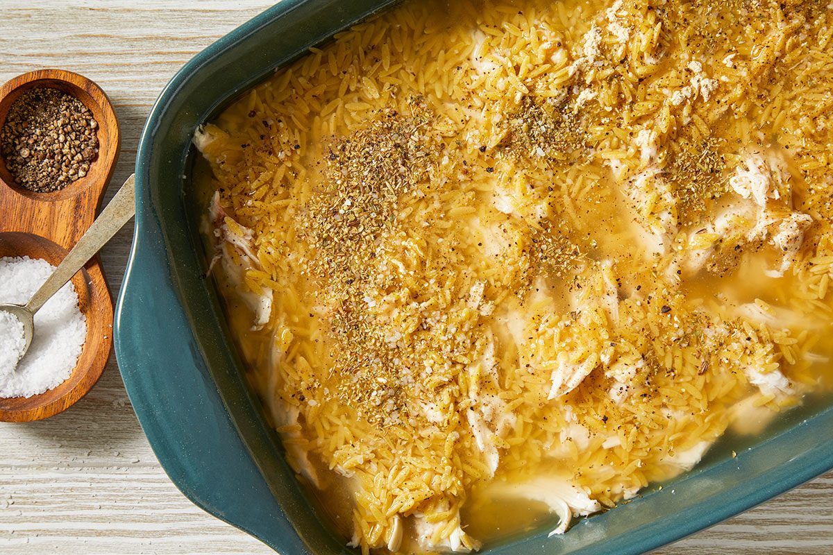 A horizontal overhead image shows a casserole dish filled with orzo, shredded chicken, broth, and seasoning sprinkled across the surface before baking.