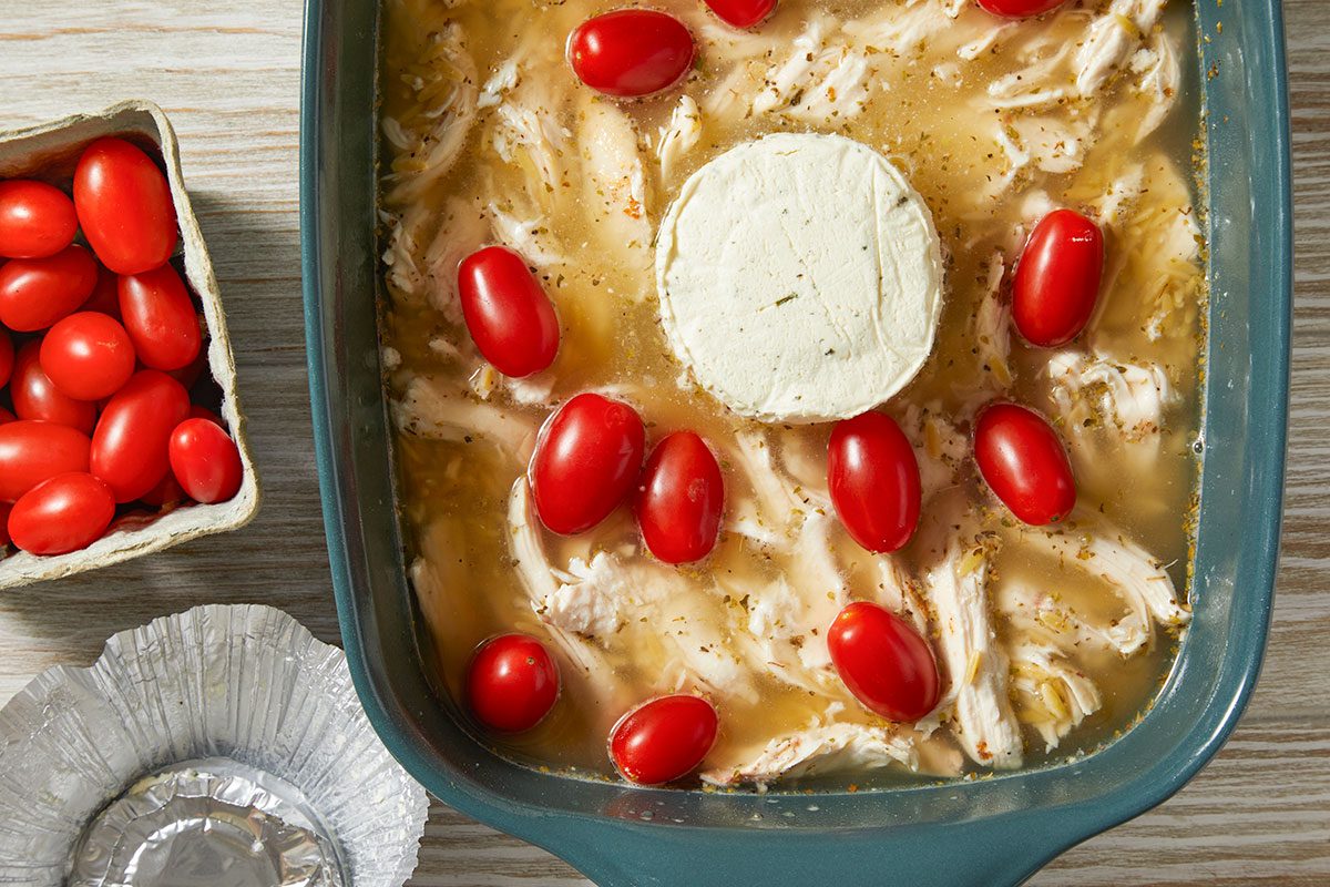 A horizontal overhead image shows a casserole dish with a round of Boursin cheese placed in the center, surrounded by grape tomatoes and uncooked orzo mixture.