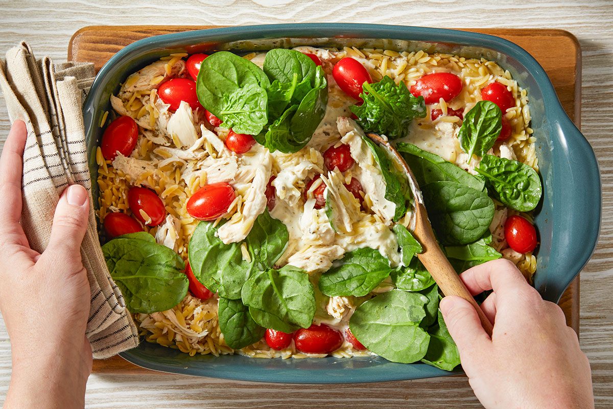 A horizontal overhead image shows fresh baby spinach being added to a casserole dish filled with orzo, chicken, and tomatoes, with hands mixing the ingredients.