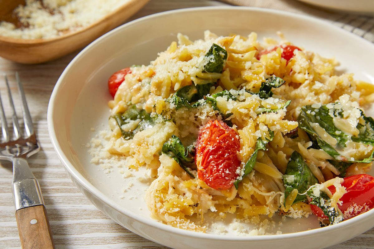 A horizontal close-up image shows a serving of chicken orzo bake in a bowl with visible spinach, tomatoes, and grated Parmesan cheese.
