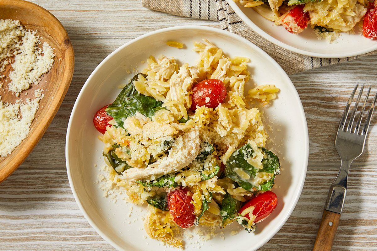 A horizontal overhead image shows two plates of chicken orzo bake with visible spinach, tomatoes, and grated cheese, arranged on a tabletop with utensils.
