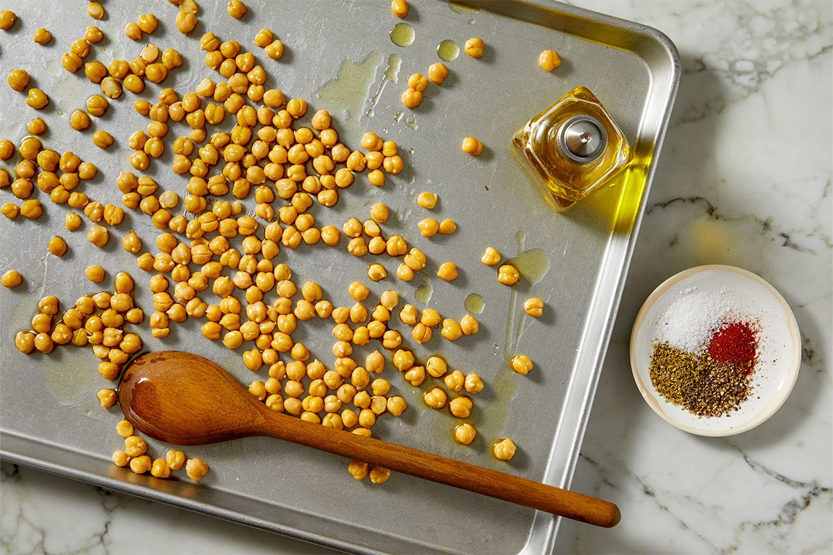 Overhead horizontal image of chickpeas being tossed with a wooden spoon on a baking sheet. Olive oil, salt, pepper, oregano and paprika are visible ingredients on a marble countertop.