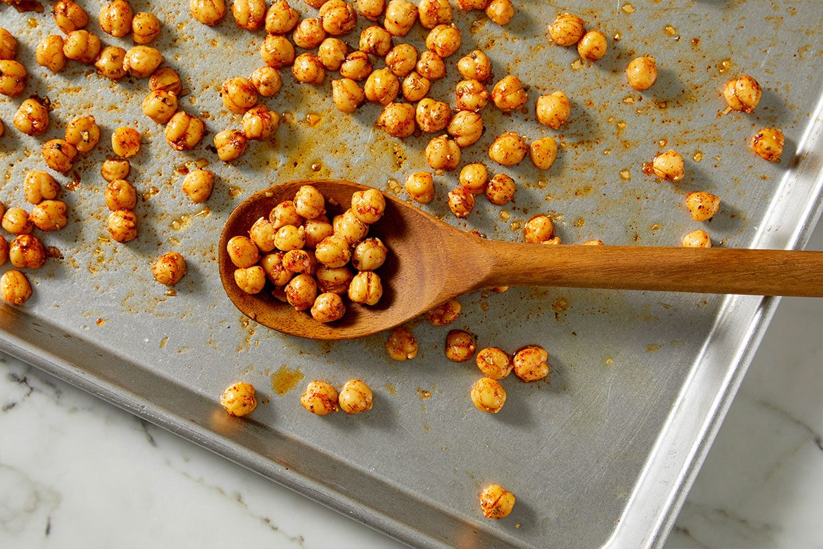 Overhead horizontal image of spiced chickpeas spread across a baking sheet with a wooden spoon holding some. The chickpeas are golden brown and covered in spices, sitting on a metal baking sheet.