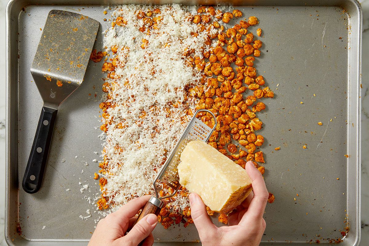 Overhead horizontal image of hands grating cheese over caramelized corn kernels on a sheet pan. The grated cheese is piling up on top of the corn, a metal spatula is resting nearby and a grater can be seen in the hands.