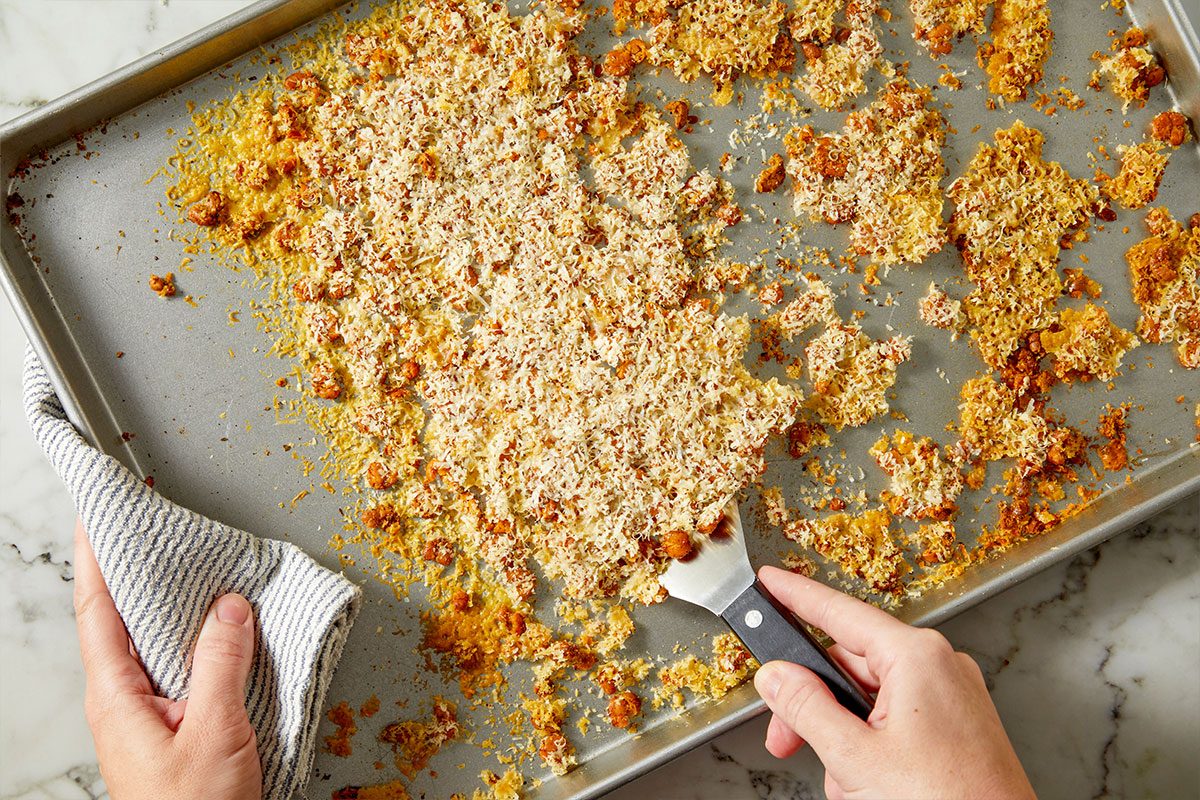 Overhead horizontal image of chickpeas and cheese on a baking sheet. A hand holds a metal spatula, separating the baked chickpeas and cheese on the tray.