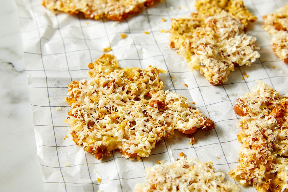 Overhead horizontal image of golden brown fried food on white parchment paper with grey graph lines and shredded coconut. The fried food appears to be sweet or savory, crispy and flaky, and sits next to others.