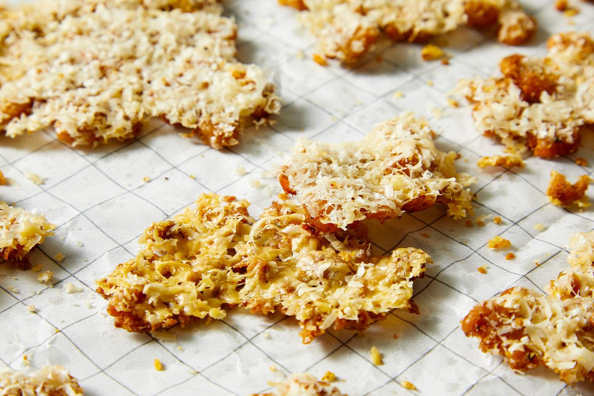 Overhead horizontal image of golden-brown fried chicken skin pieces topped with shredded Parmesan cheese on parchment paper. The image depicts a close-up view of the individual chicken skin pieces, showcasing the texture of the fried skin and cheese.