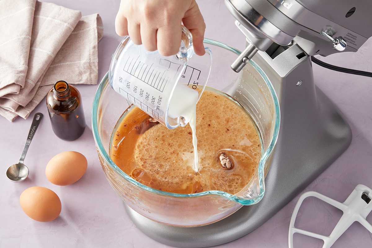 A hand pours milk from a measuring cup into a mixing bowl with eggs and other ingredients next to a stand mixer, with two eggs, a measuring spoon, and a bottle nearby on a countertop.