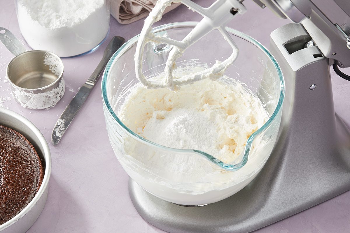 A stand mixer with a glass bowl containing white frosting is shown on a counter. Nearby are a bowl of cocoa powder, a metal measuring cup, a spoon, and a container of powdered sugar.