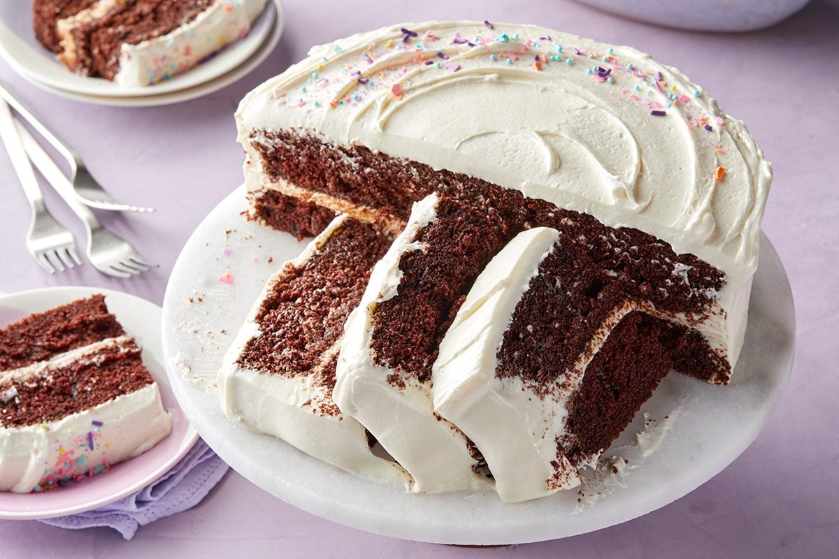 A chocolate layer cake with white frosting and sprinkles sits on a white cake stand. Several slices have been cut and served on nearby plates with forks.
