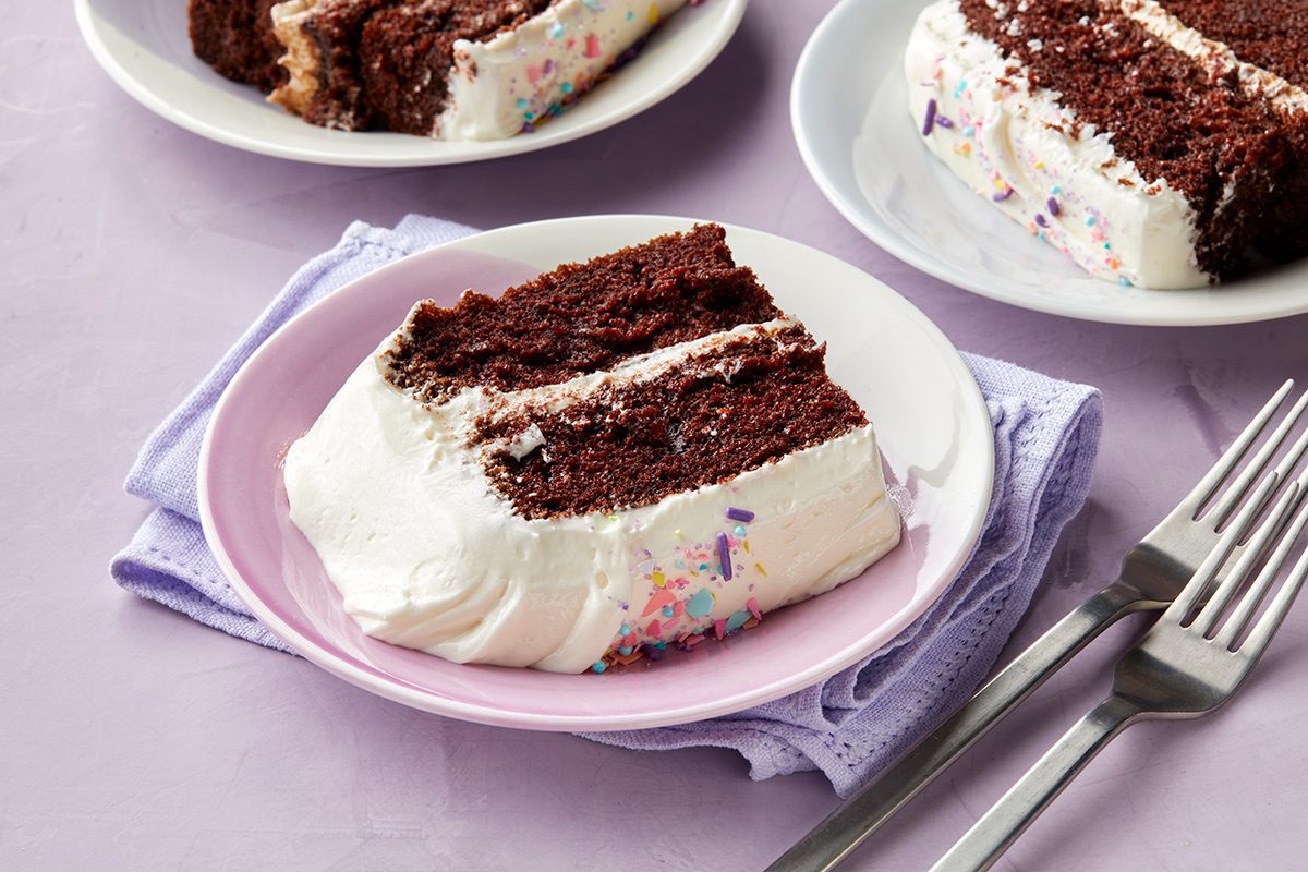 A slice of chocolate cake with white frosting and colorful sprinkles on a pink plate, next to two forks. Two more slices are visible in the background on white plates.