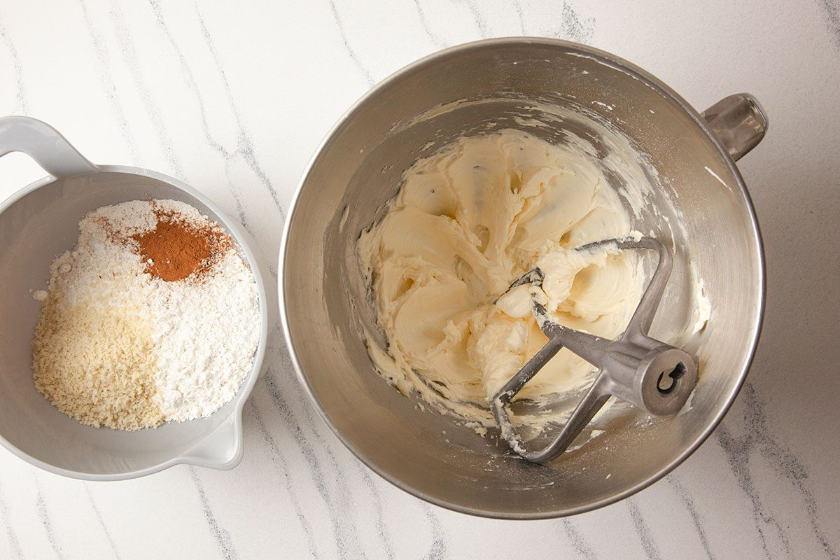 A horizontal overhead image shows a stand mixer bowl with creamed butter beside a bowl of dry ingredients including flour, panko crumbs, cinnamon, and sugar on a marble surface.