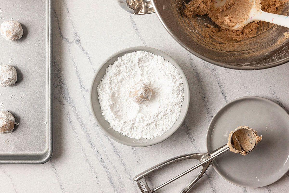 A horizontal overhead image shows cookie dough being scooped beside a bowl of powdered sugar with baking tools arranged on a clean marble surface.