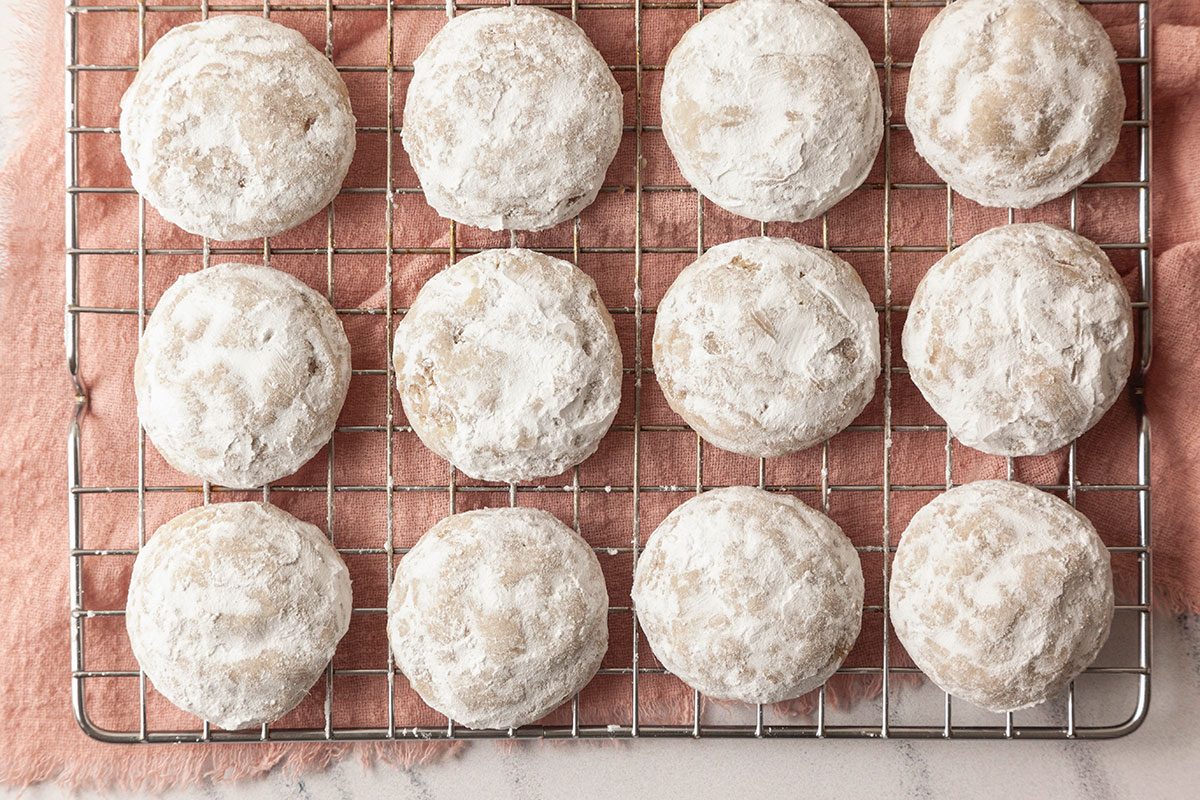 A horizontal overhead image shows baked cookies heavily coated in powdered sugar arranged on a cooling rack over a soft textile surface.