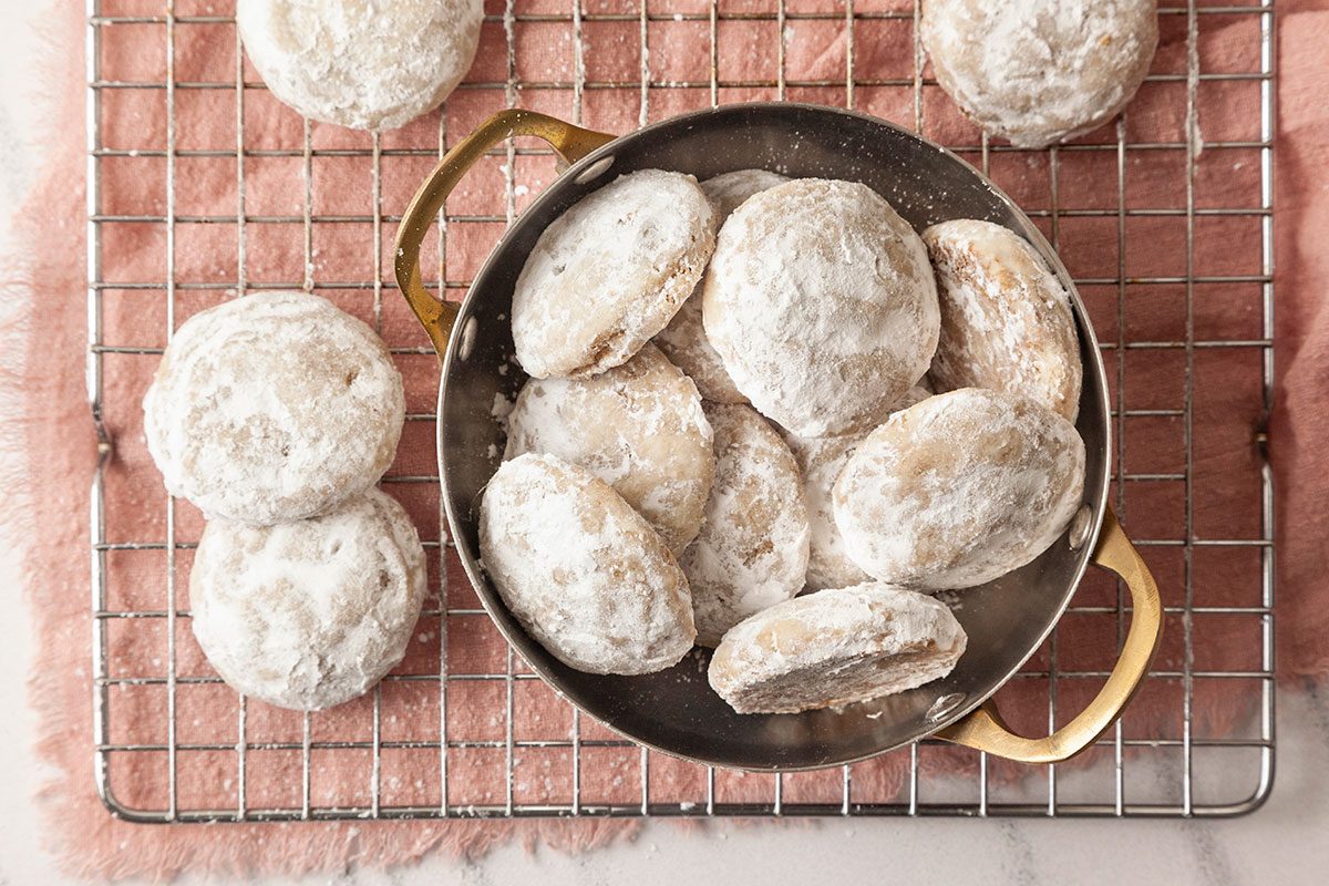 A horizontal overhead image shows powdered sugar cookies arranged in a small pan on a cooling rack with a soft fabric background and baking props.