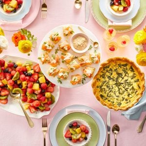 overhead styled scene shows a pastel-toned brunch table with multiple plated dishes including fruit salad, quiche, desserts, appetizers, and sparkling beverages arranged on a light surface.