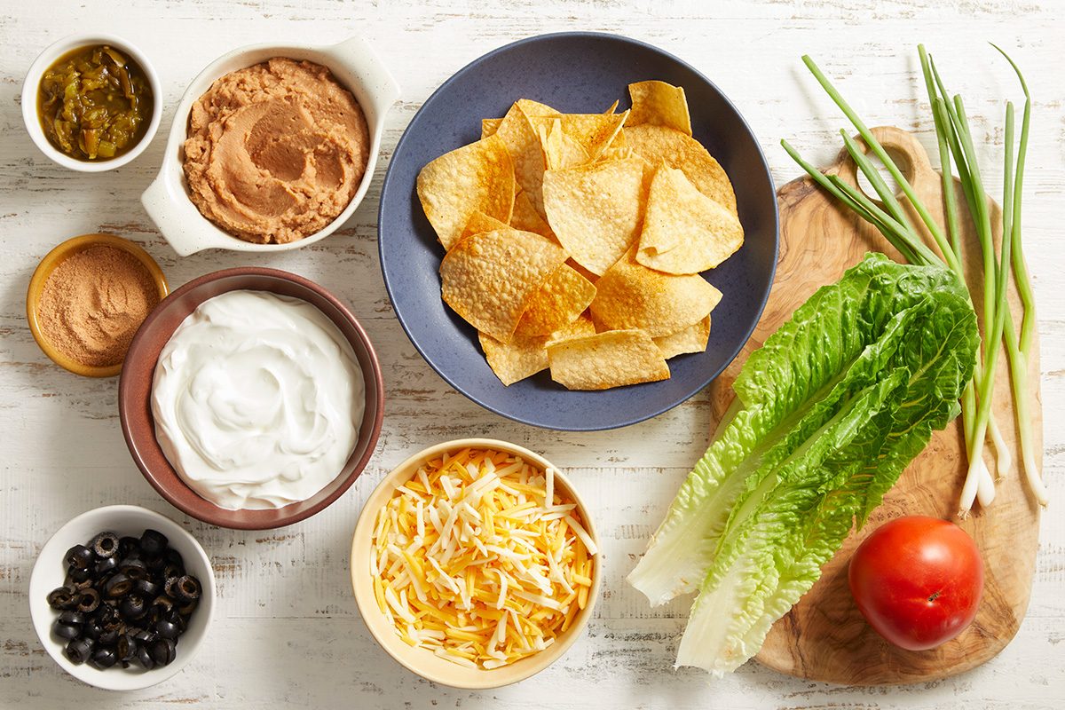 A top-down view of nacho ingredients: tortilla chips, shredded cheese, refried beans, salsa, sour cream, sliced black olives, green onions, a head of lettuce, and a whole tomato on a white surface.