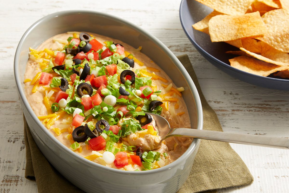 Oval dish filled with layered dip topped with shredded cheese, diced tomatoes, black olives, green onions, and lettuce, with a spoon inside. A bowl of tortilla chips sits nearby on the table.