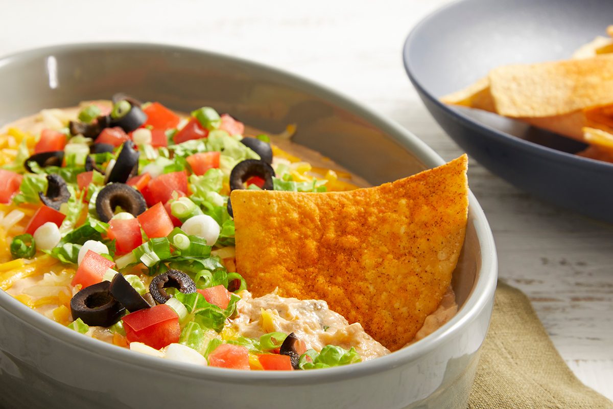 A bowl of layered dip topped with chopped tomatoes, black olives, green onions, and lettuce, with a tortilla chip dipped in. A bowl of more tortilla chips is in the background.