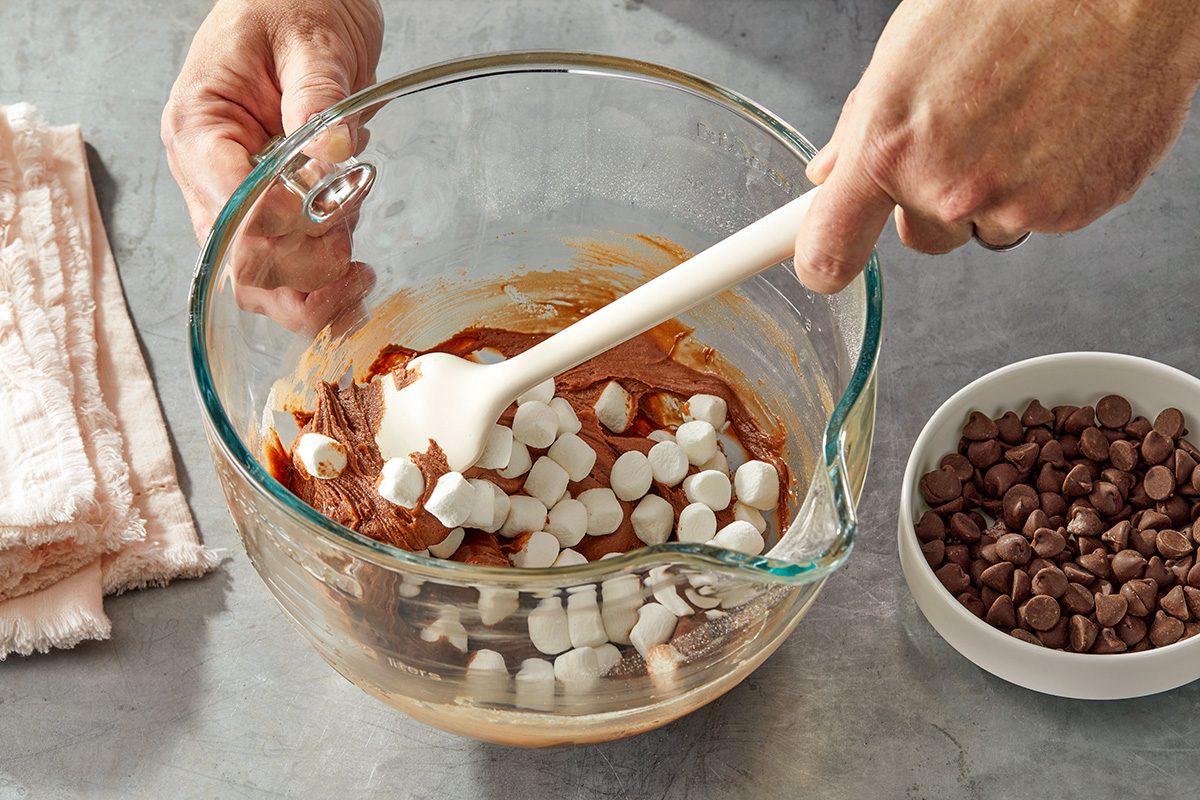 a mixing bowl where a hand is using a spatula to stir in some marshmallows into a chocolate batter