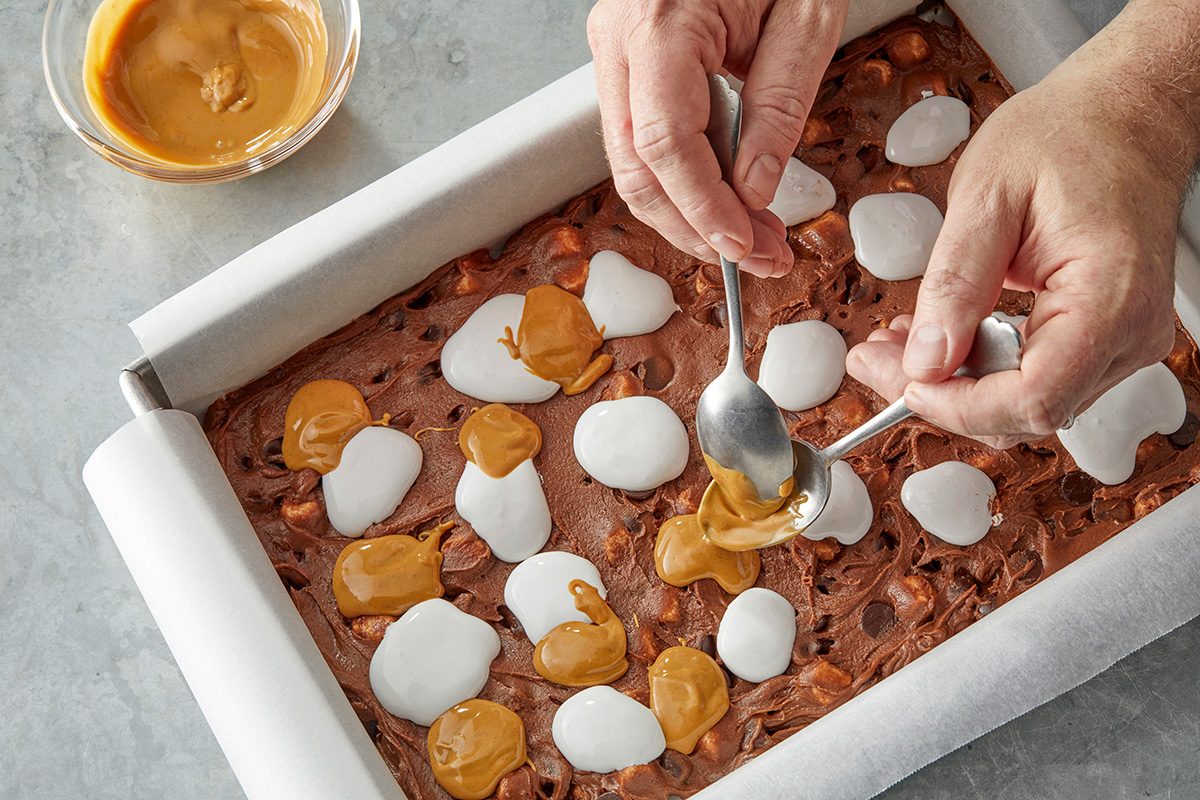 spooning caramel onto a chocolate dessert with marshmallows in a baking pan