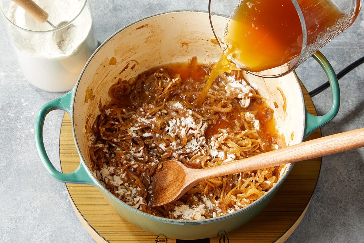 Overhead horizontal image of broth being poured into caramelized onions with flour to build the stew base. Sprinkle with flour. Add beef broth, stirring to loosen browned bits from pan. Transfer to slow cooker.