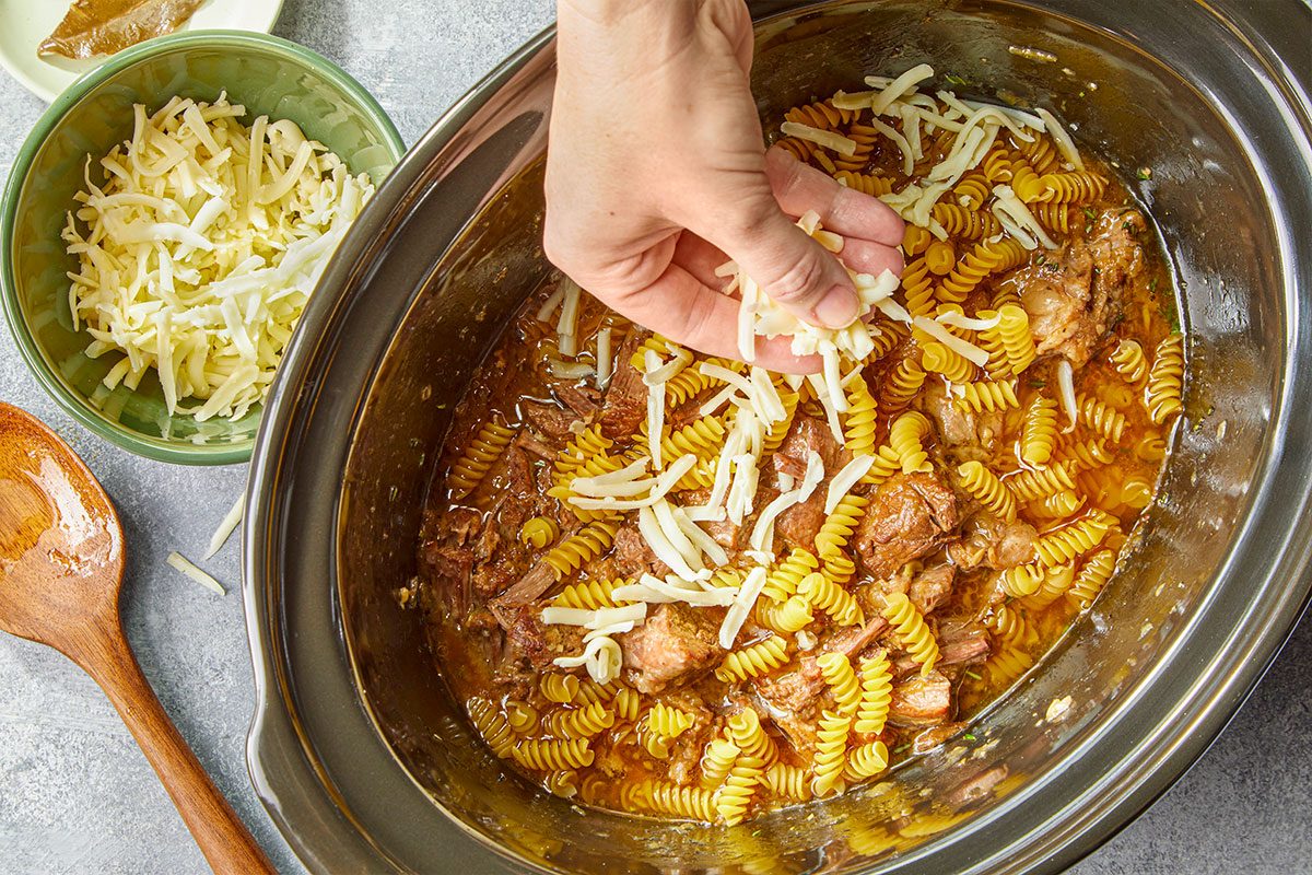 Overhead horizontal image of slow cooker filled with stew, pasta, shredded beef, and cheese being added. Add rosemary and bay leaf to slow cooker. Cover and cook on high until beef is tender, about 6 hours. Remove beef and shred with two forks. Return beef to slow cooker. Remove bay leaf; discard. Add noodles; stir to coat. Top with Monterey Jack cheese. Cook until noodles are done, 20-25 minutes longer.