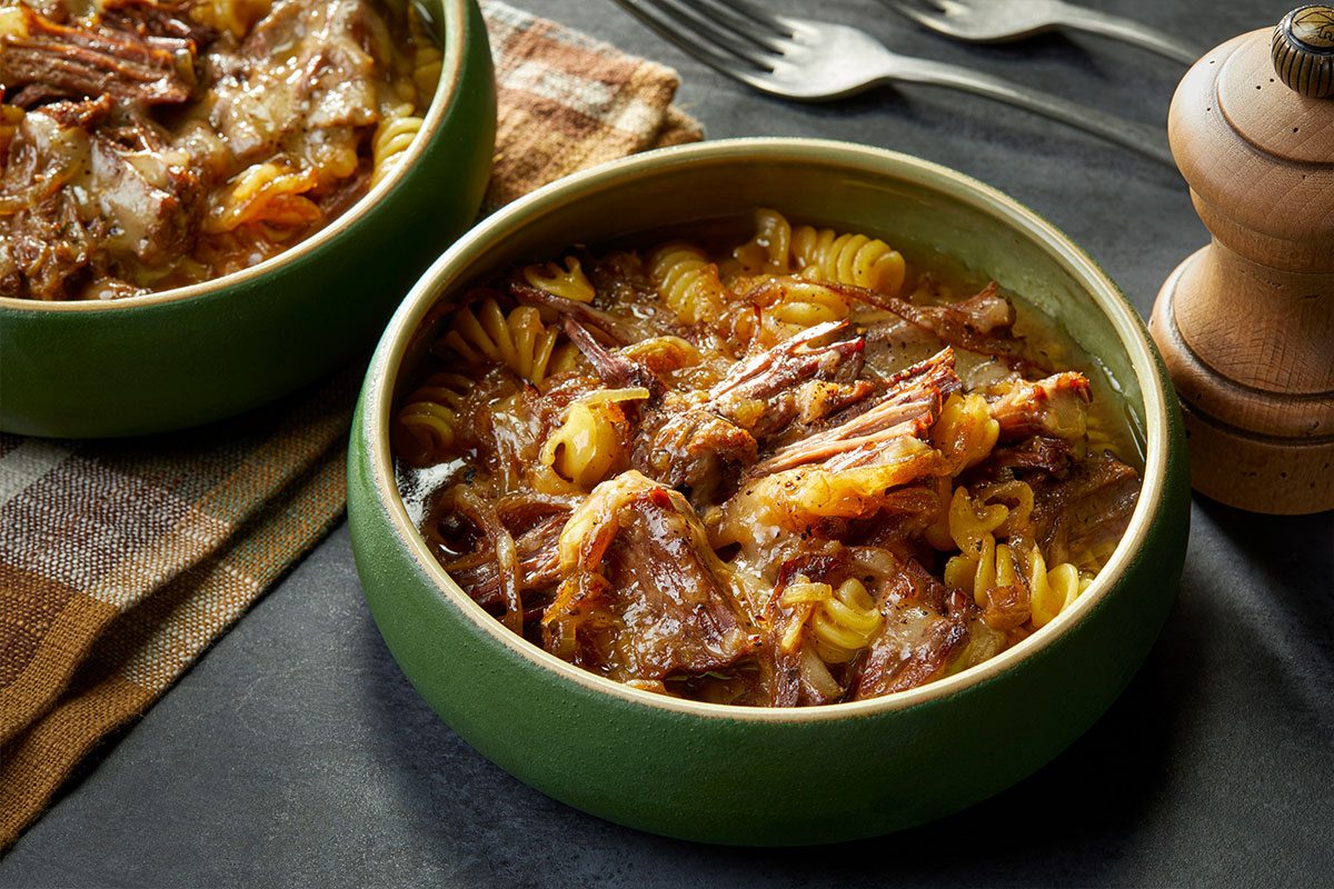 Eye-level horizontal image of a bowl of French onion stew with tender shredded beef, pasta, and melted cheese.