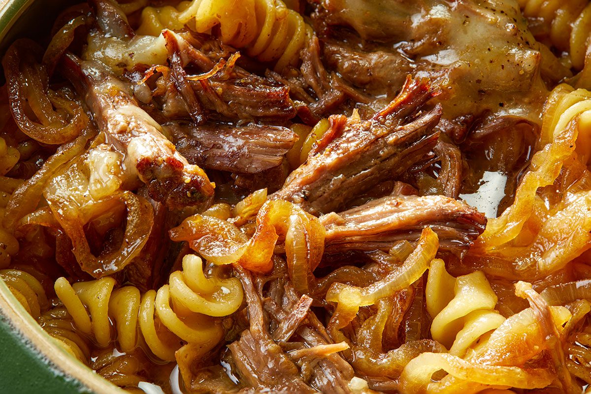 Eye-level horizontal close-up of French onion stew highlighting tender beef, caramelized onions, pasta, and melted cheese.