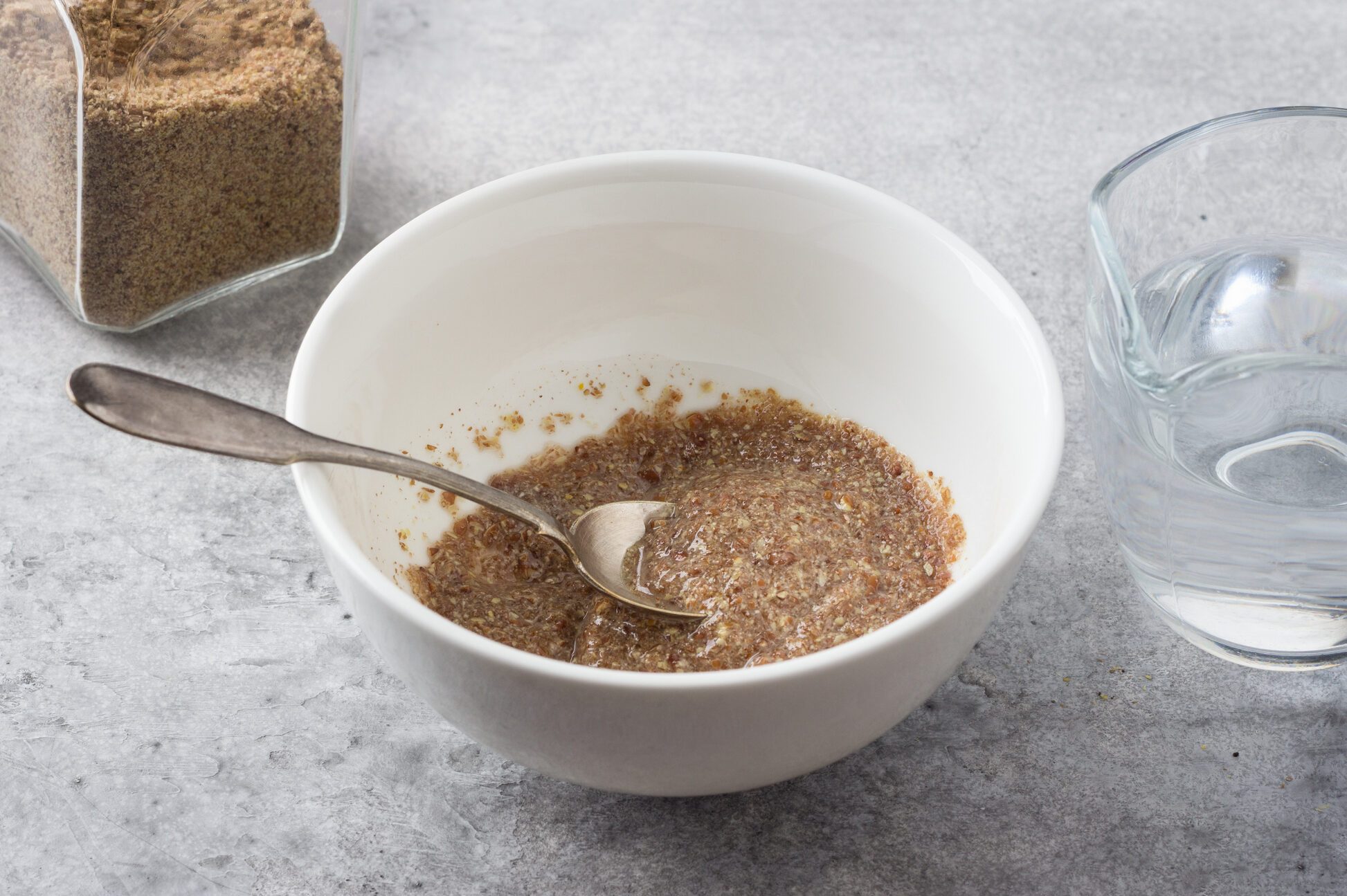 A bowl with ground flax and water, a jar of ground flax and a jug of water on a gray textured background. Cooking a vegan flax egg.