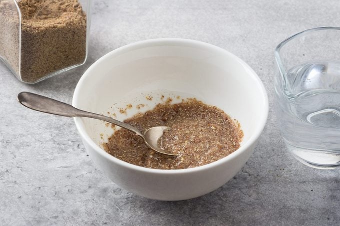 A bowl with ground flax and water, a jar of ground flax and a jug of water on a gray textured background. Cooking a vegan flax egg.