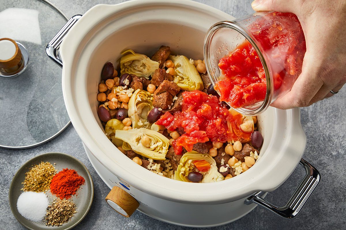 Overhead horizontal image of diced tomatoes being added to a slow cooker with lamb, chickpeas, olives, and artichokes.