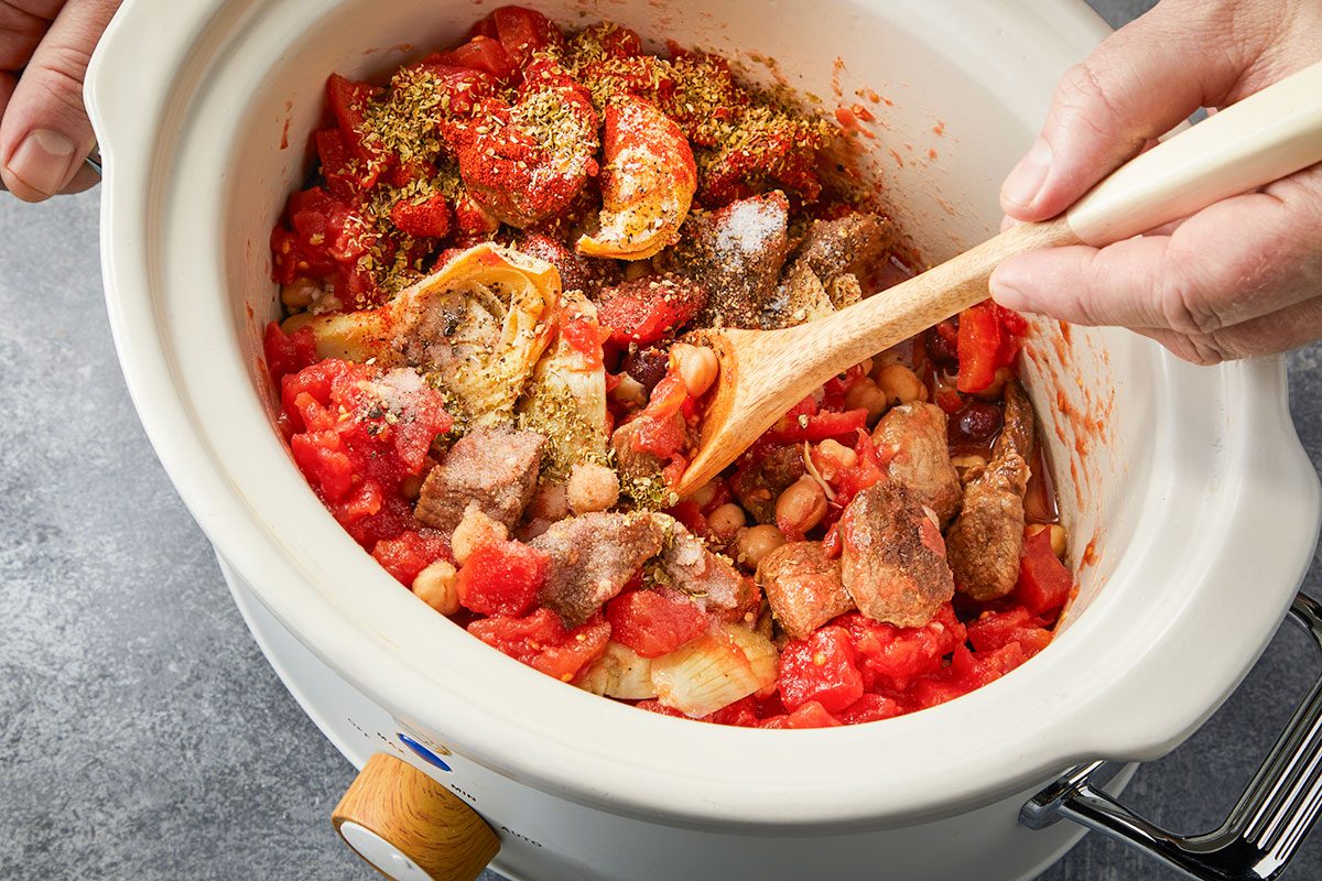 Overhead horizontal image of the lamb stew mixture being stirred in a slow cooker with tomatoes, spices, and vegetables.