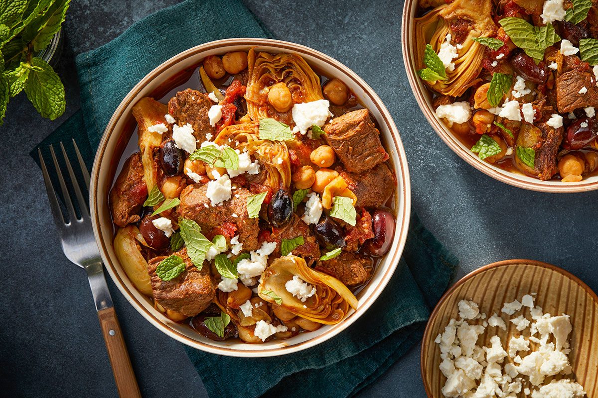 Overhead horizontal image of a serving bowl of Greek style lamb stew with feta cheese, olives, and herbs.