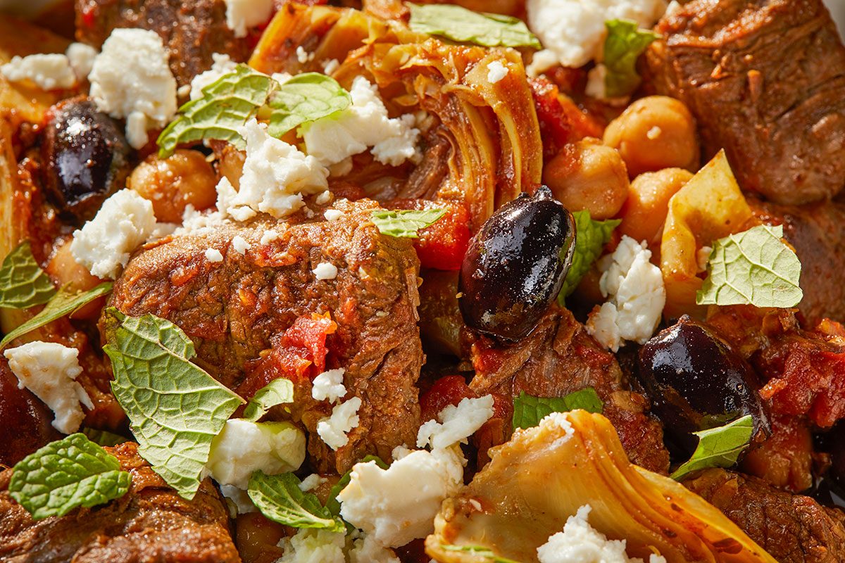 Overhead horizontal close-up of Greek style lamb stew showing tender lamb, chickpeas, olives, feta, and herbs.