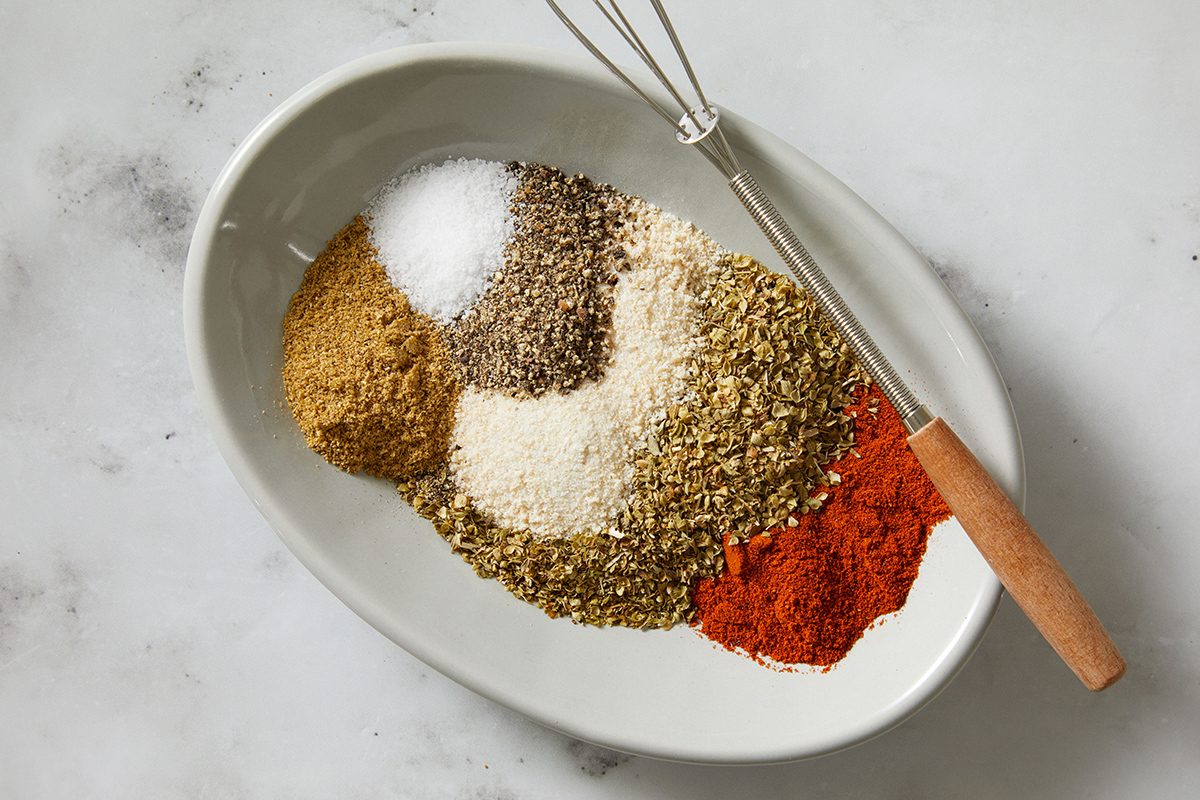An oval white plate holds separate piles of various spices, including salt, pepper, paprika, onion powder, and others, next to a small whisk with a wooden handle on a light surface.