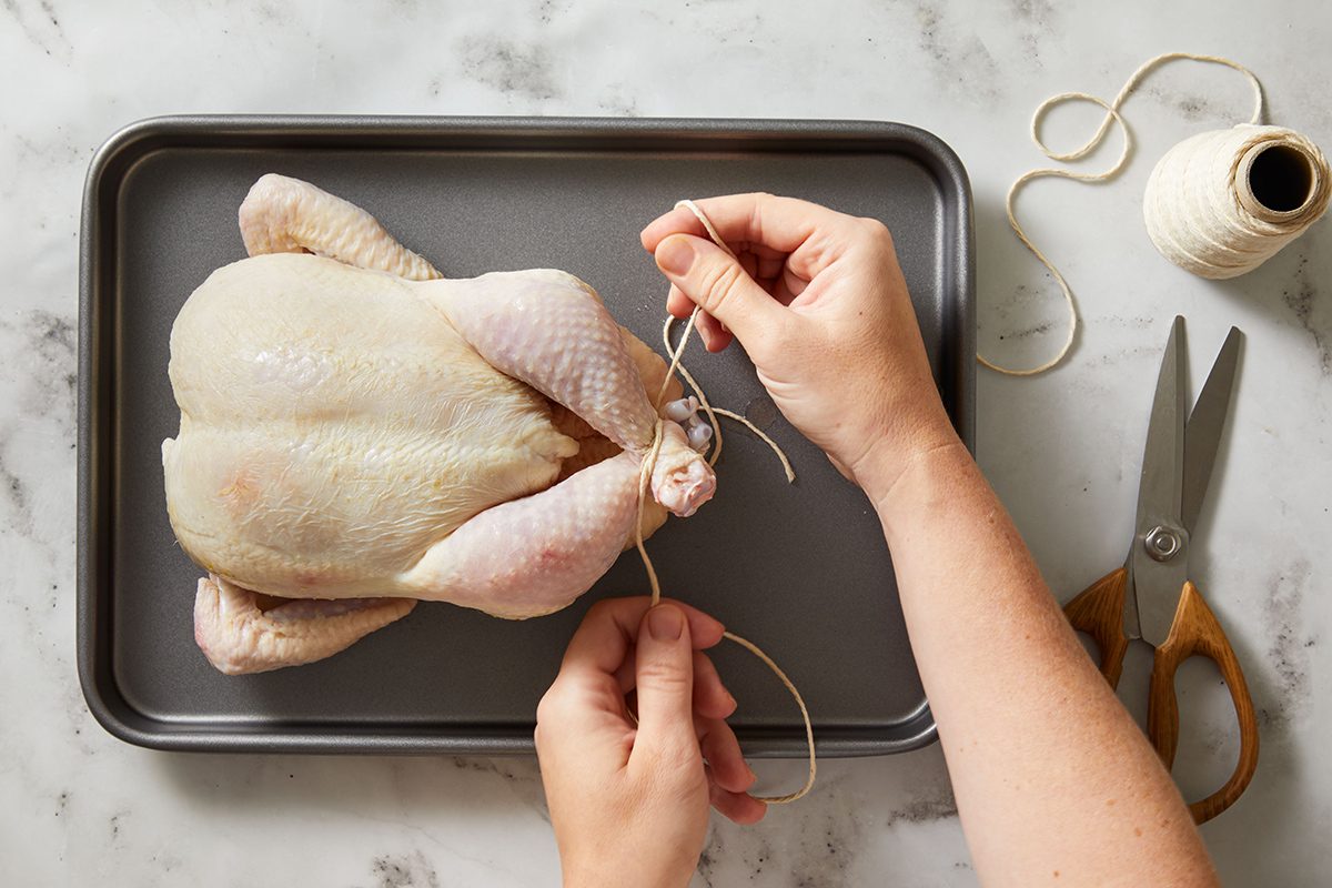 A person ties butcher’s twine around the legs of a raw whole chicken on a baking tray, with scissors and a spool of twine nearby on a marble countertop.