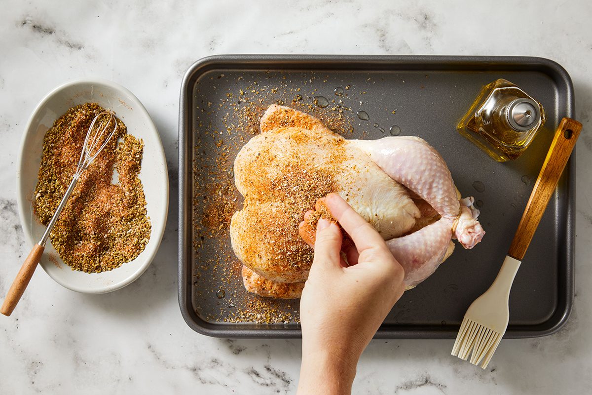A hand sprinkles seasoning onto a raw whole chicken on a baking tray. Nearby are a bowl of spice mix with a fork, a bottle of oil, and a basting brush, all on a marble countertop.