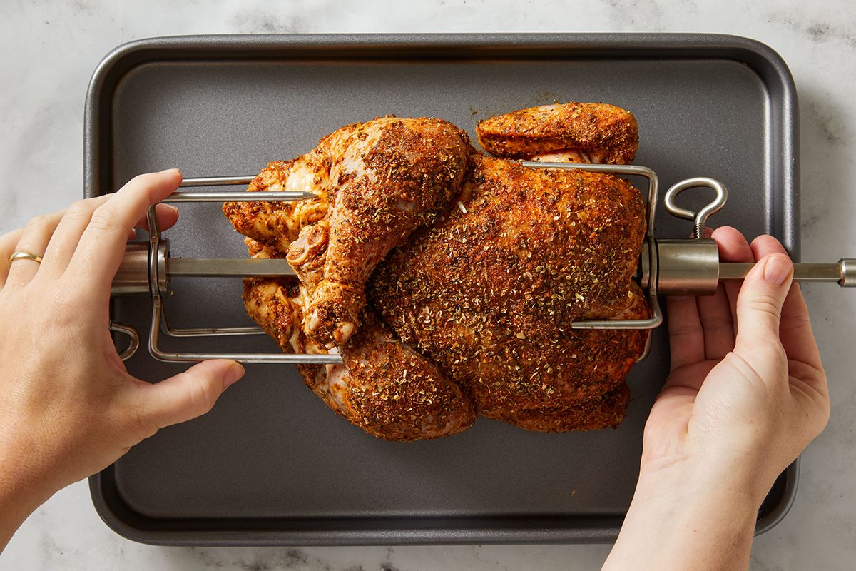 Hands securing a seasoned whole chicken onto a metal rotisserie spit over a baking tray, preparing it for roasting.