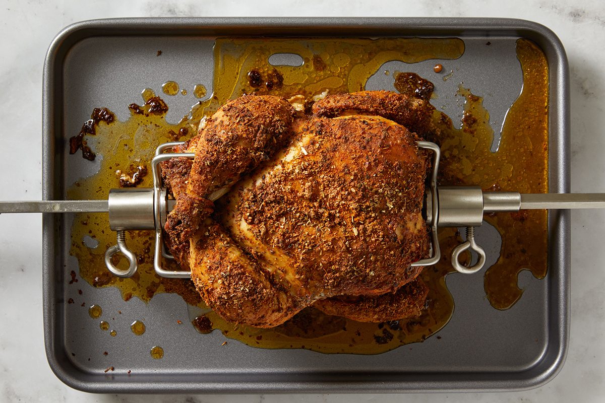 Whole rotisserie chicken with crispy, seasoned skin is mounted on a metal spit and placed over a baking tray, with juices and spices collected below.