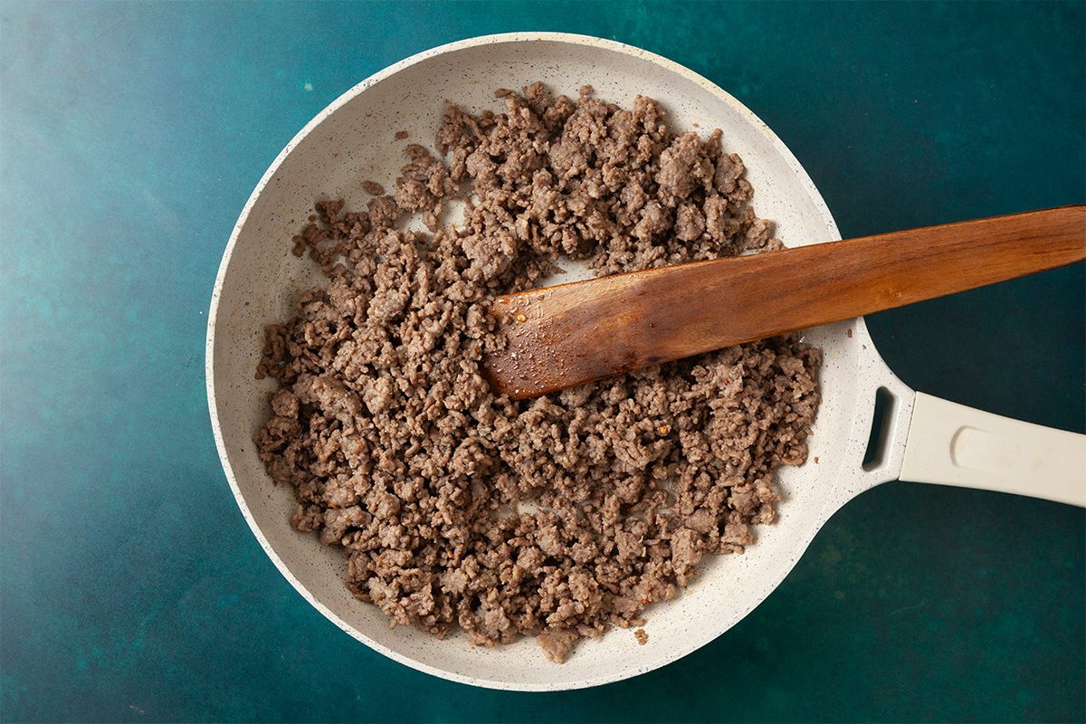 Overhead horizontal image of ground beef and sausage browning in a skillet with a wooden spoon.