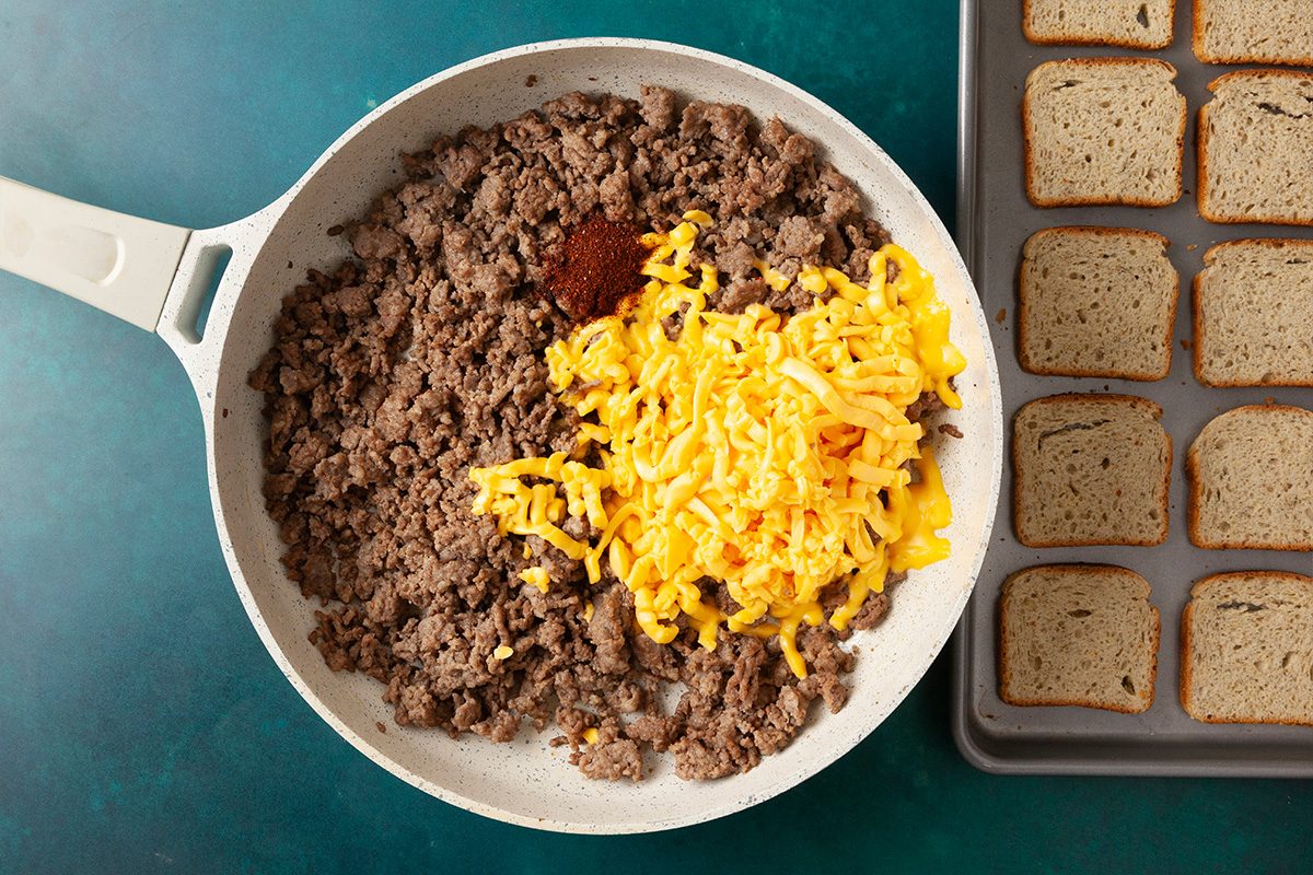 Overhead horizontal image of browned meat mixture combined with shredded cheese in a skillet next to sliced rye bread.