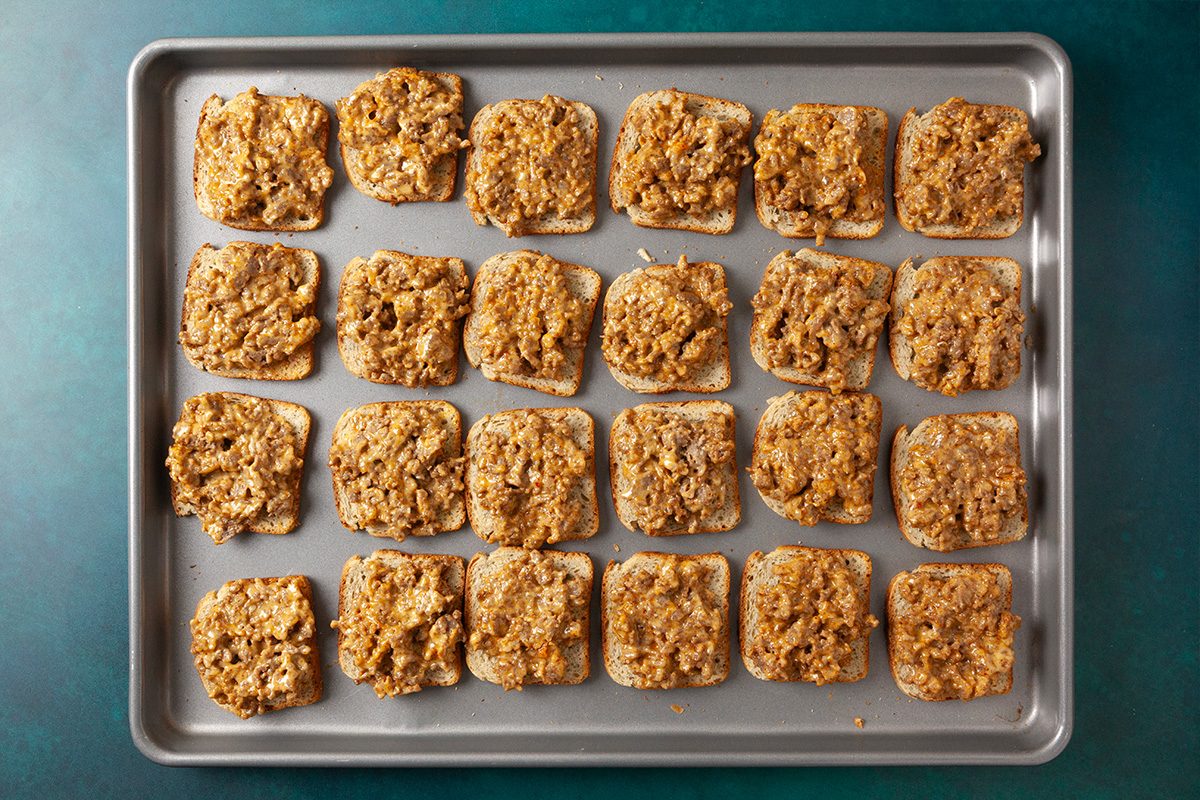 Overhead horizontal image of rye bread slices topped with meat and cheese mixture arranged on a baking sheet.