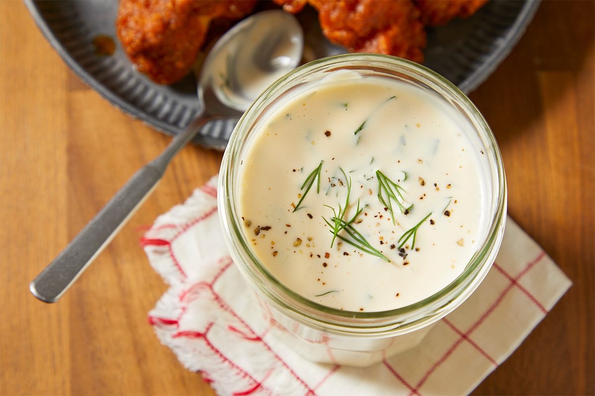 A horizontal overhead image shows a jar of creamy ranch dressing topped with fresh dill and seasoning with a spoon and fried food in the background.